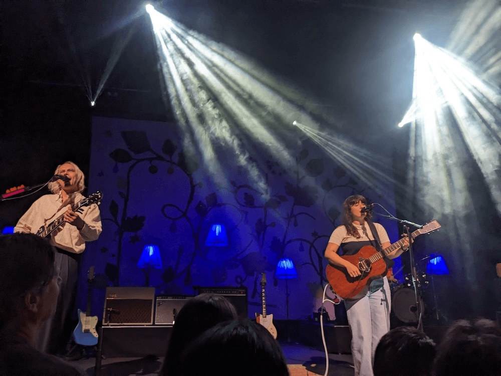 Liz, an Indonesian-pakeha woman wearing jeans and a tshirt and holding a beautiful acoustic guitar, stands next to Jono, a blond man with long hair and an electric guitar, with white lights and a blue background