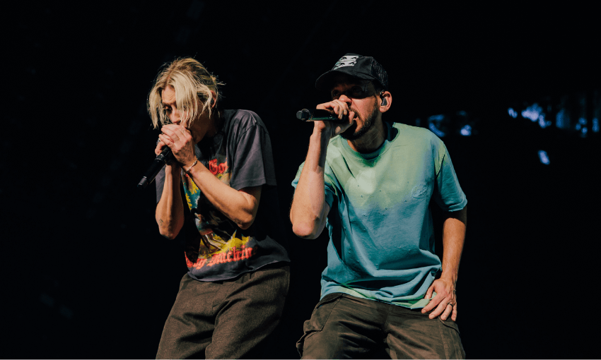 Two musicians performing on stage, both holding microphones. One wears a graphic t-shirt and has blond hair, while the other wears a cap and a plain t-shirt. The stage background is dark.