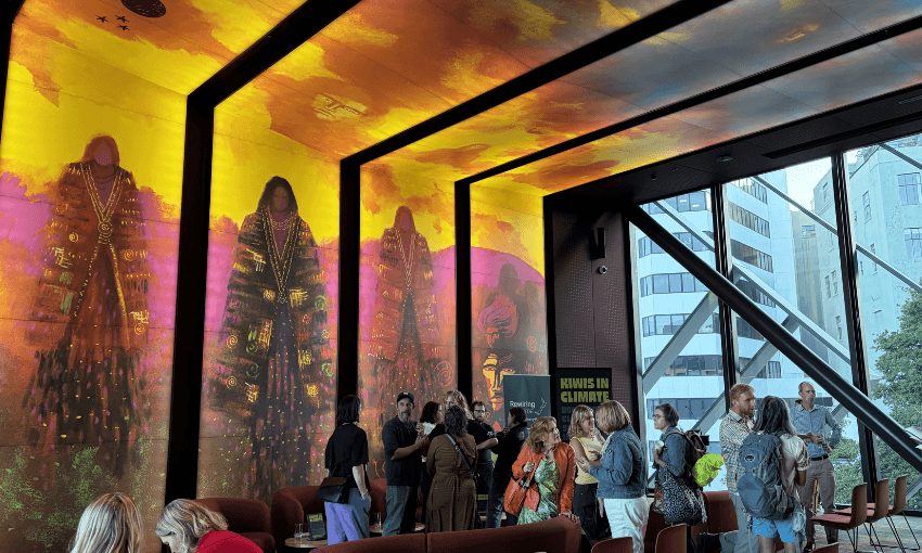 A crowd mills around speakers at the front of stunning room adorned with wall to ceiling artwork of earth mothers.