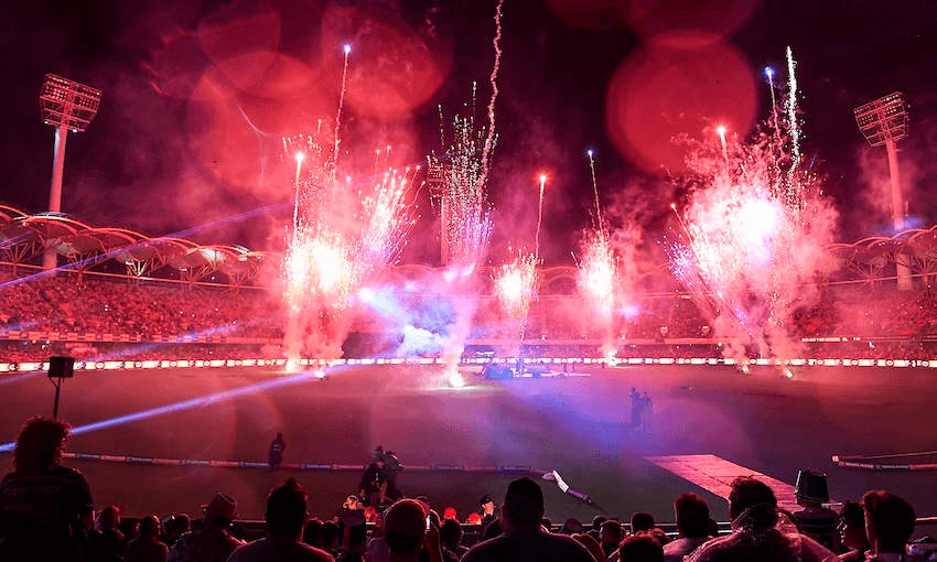 Fireworks light up the night sky over a crowded sports stadium, with bright red flares and smoke above the cheering audience and illuminated field.