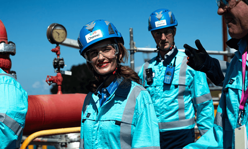 A group of people in blue safety gear and helmets stand outdoors near industrial piping and gauges. A woman in front is smiling at the camera, while others are engaged in conversation.