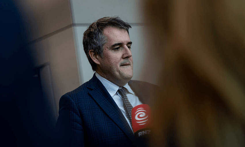 A man in a suit and tie stands indoors, holding a red RNZ microphone. He appears to be speaking or preparing to speak, with blurred figures and a wall in the background.