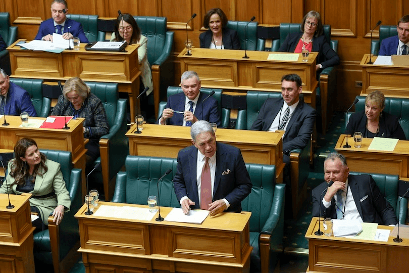 A man in a suit speaks at a podium in a government chamber, surrounded by seated officials and members of parliament listening and observing. Desks with papers, glasses of water, and microphones are visible.