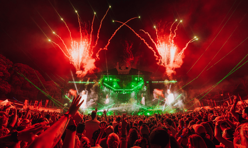 A large crowd at a nighttime outdoor music festival raises their hands as green lights and red fireworks illuminate the stage. The atmosphere is energetic and festive.