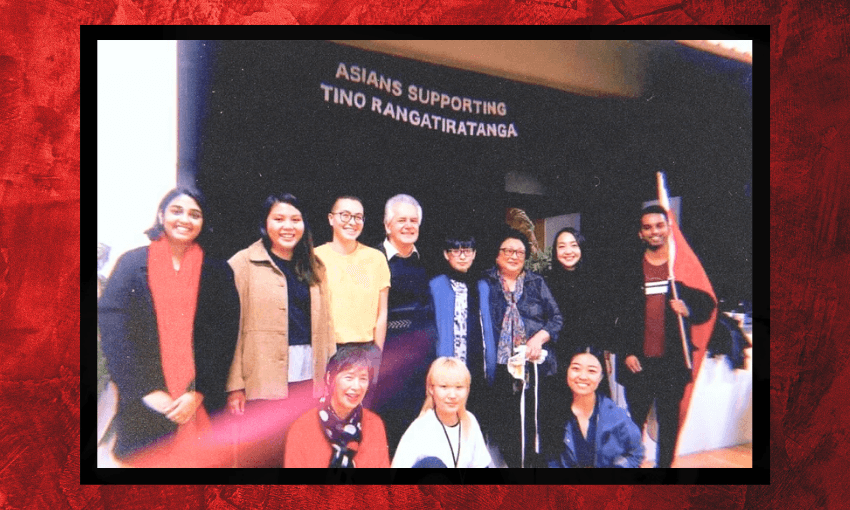 A group of people smiling and posing together in front of a sign that reads "ASIANS SUPPORTING TINO RANGATIRATANGA." One person holds a flag with a red, black, and white design.