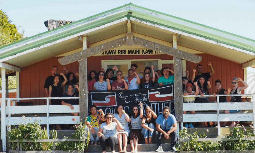 A large, diverse group of people pose together on the steps and porch of a wooden building with a green roof, holding a black banner with white and red designs. Some people have fists raised; everyone is smiling.