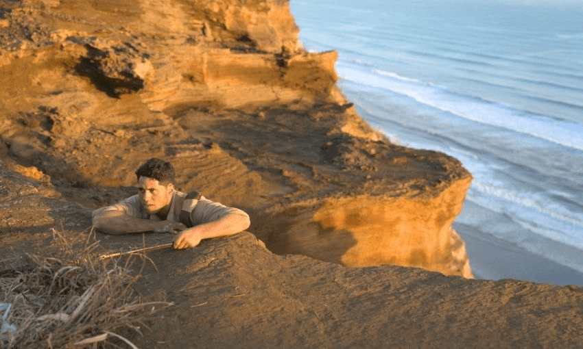 A man in a brown shirt clings to the edge of a rocky cliff overlooking the ocean, with waves gently rolling onto the beach far below.