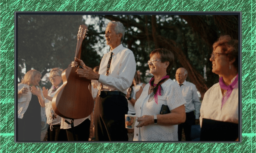 A group of older adults, some holding musical instruments and mugs, stand outdoors under trees. They wear white shirts, black pants, and purple scarves, smiling and appearing ready to perform.