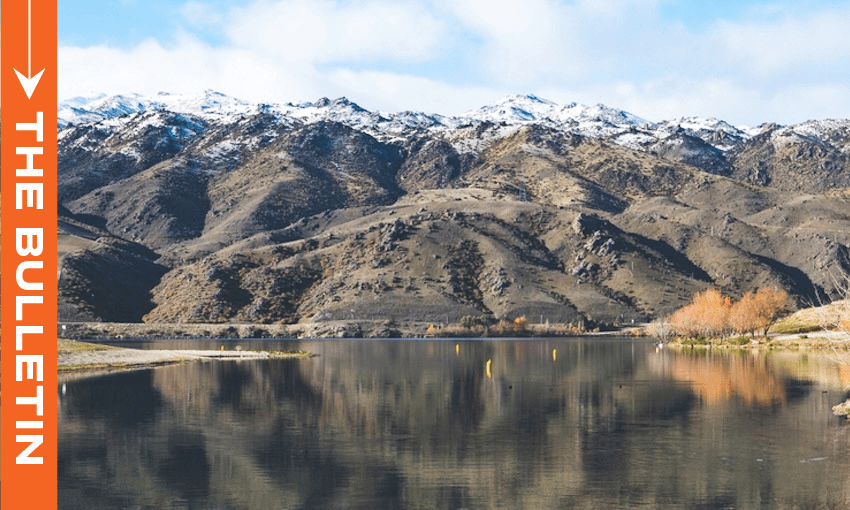 A calm lake with clear reflections sits in front of rugged, snow-dusted mountains under a partly cloudy sky. On the left, an orange vertical banner reads "THE BULLETIN" in white text.
