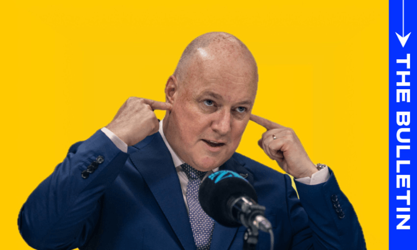 A bald man in a blue suit gestures by plugging his ears with his fingers while speaking into a microphone. The background is bright yellow, with a vertical blue banner reading "The Bulletin" on the right.