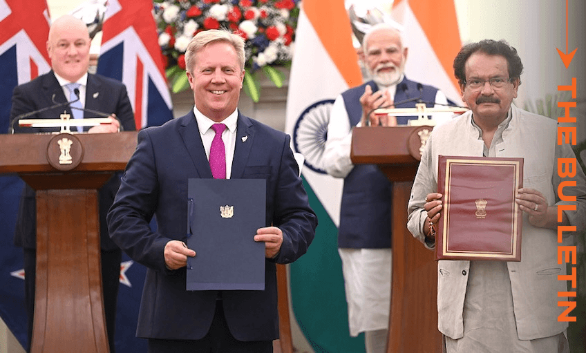 Four men stand in front of New Zealand and Indian flags. Two men in front hold official documents, while two others, including Indian PM Modi, stand behind podiums smiling during a formal signing ceremony.