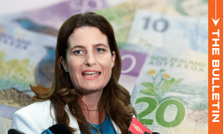 A woman speaking at a press conference, with microphones in front of her. In the background, there are New Zealand banknotes and an orange vertical banner reading "THE BULLETIN.