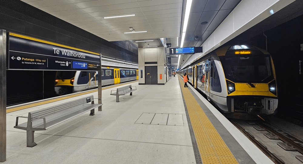 A modern underground train station with a yellow and white train on the right, benches on the platform, digital signs overhead, and a person in a safety vest standing near the train.
