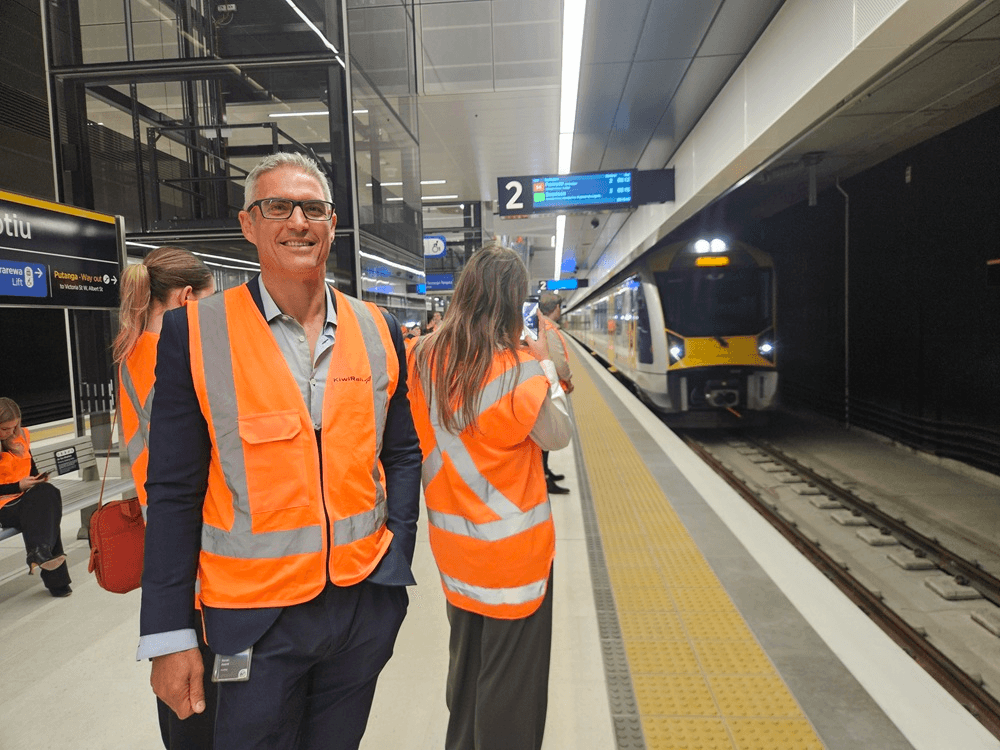 People wearing orange safety vests stand on a modern train platform as a yellow and white train arrives. One man in the foreground smiles at the camera, while others face the train. The station looks new and well-lit.