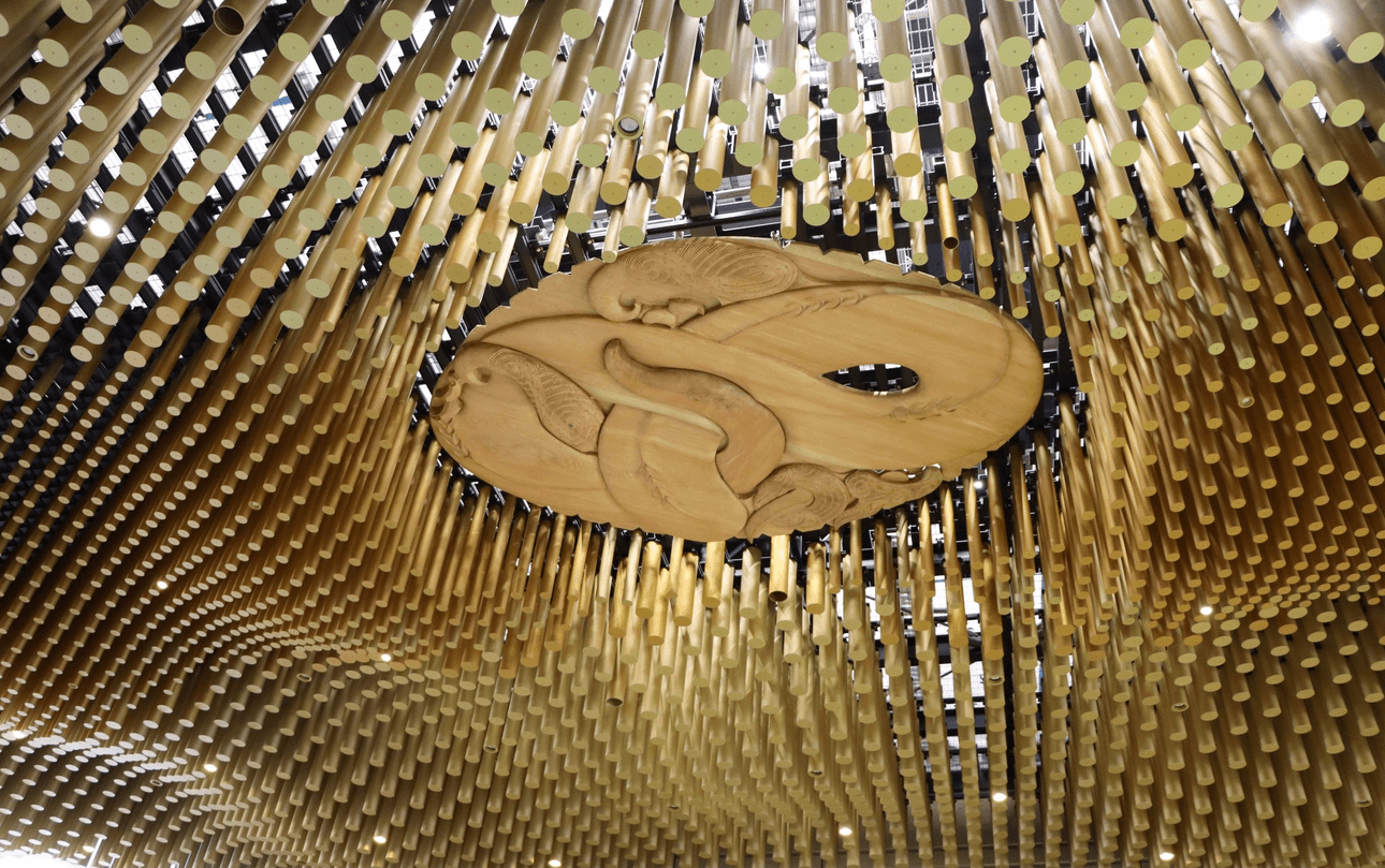Wooden ceiling art installation featuring a large central carving of two birds surrounded by golden cylindrical rods hanging down in a wavy pattern, creating an intricate and dynamic design.