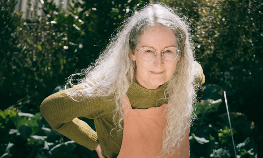 A photo of the writer Elizabeth Knox who has long grey-white hair, is wearing salmon coloured dungarees and is looking up at the camera from where she's standing outside in front of a garden. It is sunny.