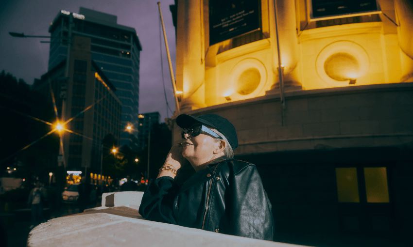 A person wearing sunglasses, a black cap, and a leather jacket smiles while leaning on a railing in a city at night, with a brightly lit building in the background.