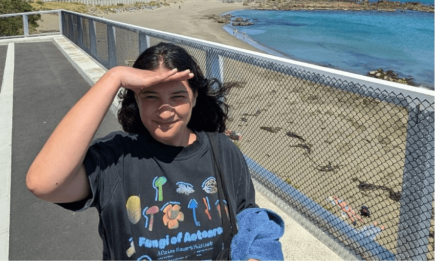 Lyric stands with her hand shading her eyes from the sun in front of a long beach.