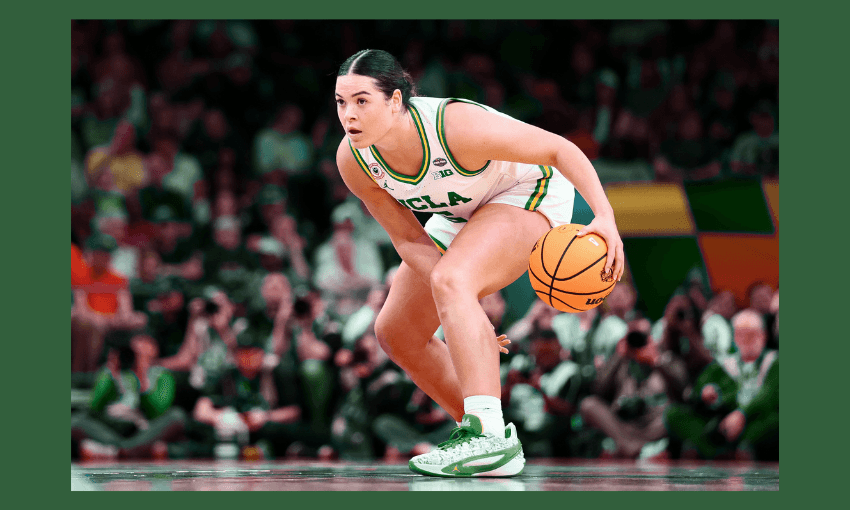A female basketball player in a UCLA uniform dribbles a basketball on the court, focused on the game, with a crowd of spectators blurred in the background.