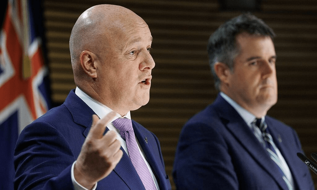 Two men in suits stand at a podium, one speaking and gesturing with his hand, the other listening. A partial view of a flag is visible in the background.