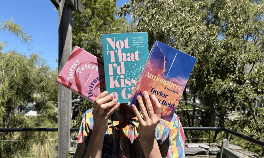 A photo of someone holding up three books in a fan formation, covering their face. They are outside and it's a sunny day.
