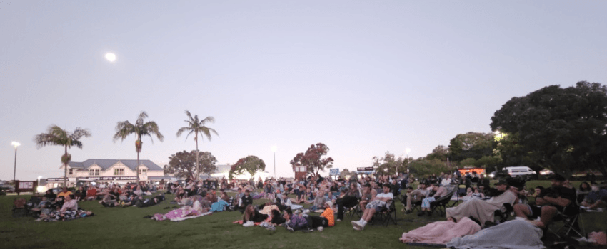 A photo of a twilight scene: people are nestled on picnic blankets on grass with palm trees in the background.