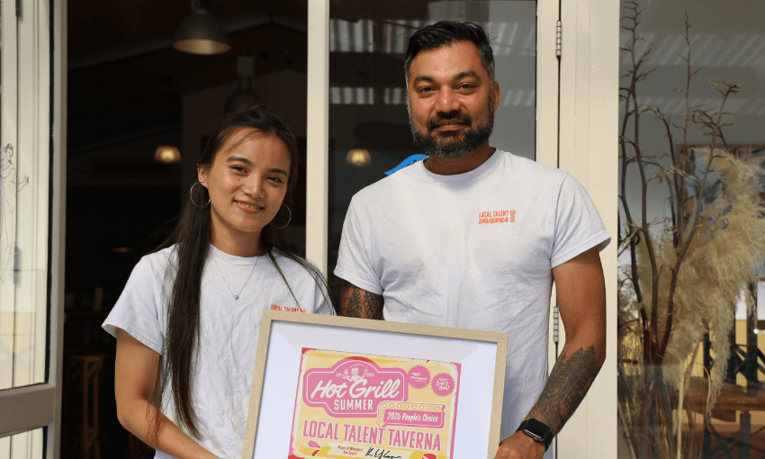 a woman and man stand outside a restaurant wearing white uniform t-shirts and holding a framed sign with the name Local Talent Taverna on it