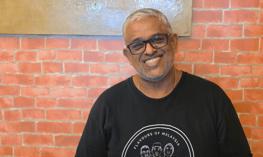 a smiling middle-aged man with brown skin, white hair and stubble, and dark-rimmed glasses