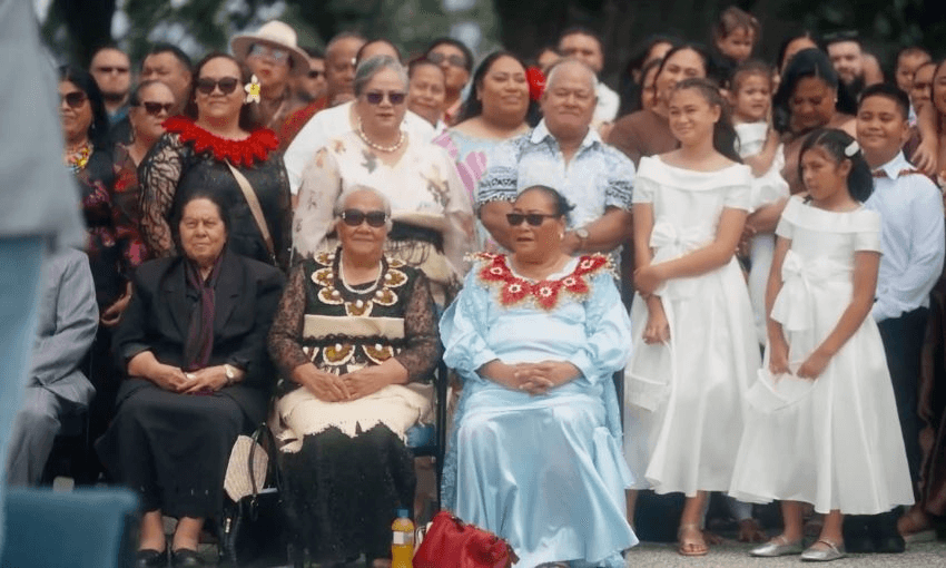 A large group of people, including women in formal dresses and traditional attire, pose together outdoors. Some are seated while others stand behind them, smiling under sunny weather.