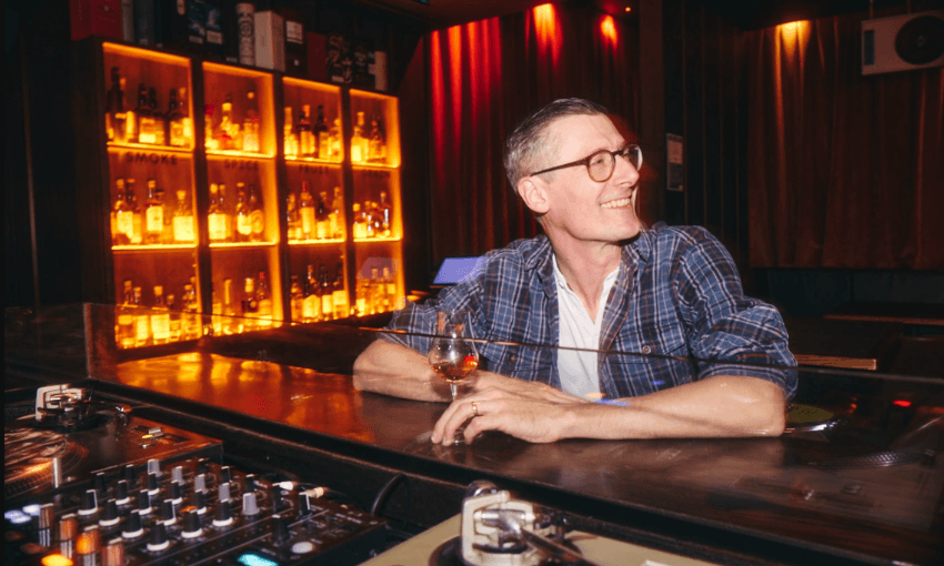 A man wearing glasses and a plaid shirt sits at a bar holding a drink, smiling. Behind him are shelves filled with bottles, softly lit by warm yellow lights. DJ equipment is visible in the foreground.