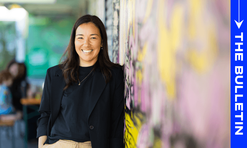 A woman with long dark hair, wearing a black blazer and tan pants, smiles while leaning against a colorful wall. The words "THE BULLETIN" appear vertically on a blue strip on the right side of the image.