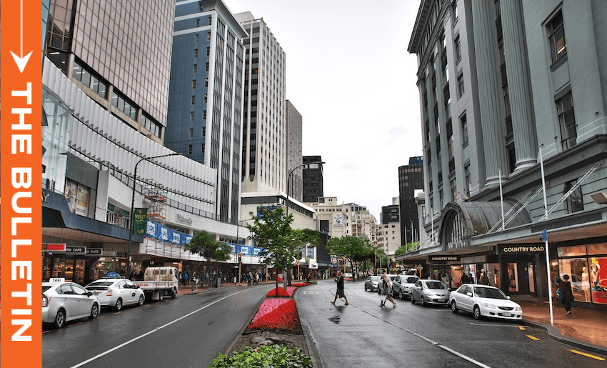 A busy city street with tall buildings, shops, cars, and people crossing the road on a cloudy day. A red and green flowerbed lines the center. An orange banner on the left reads "THE BULLETIN.