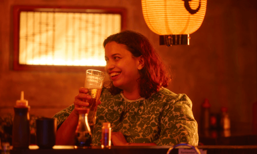 A woman in a green patterned top sits at a dimly lit bar, smiling and holding a glass of beer. Warm yellow light from a paper lantern illuminates the scene, creating a cozy atmosphere.