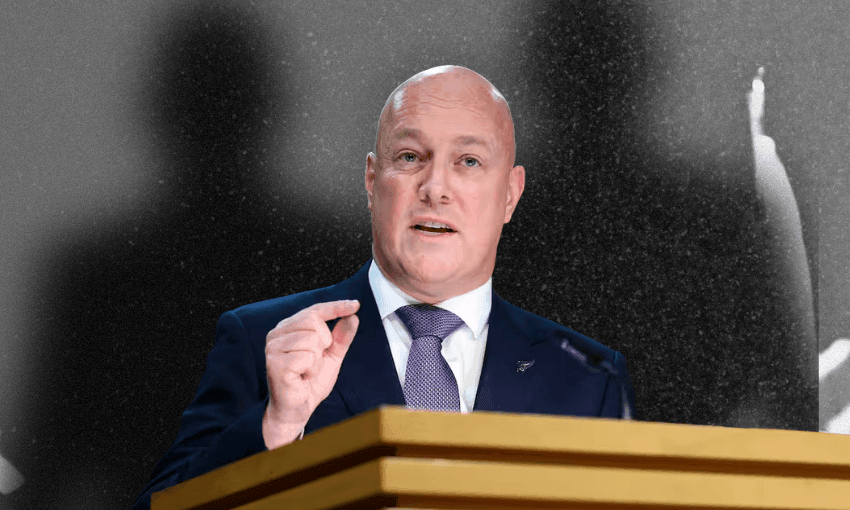 A bald man in a suit and tie speaks at a podium, gesturing with his hand. The background is black and white with blurred shadows of people.