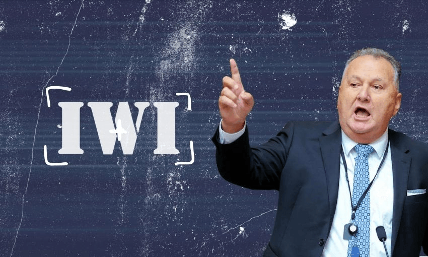 A man in a suit gestures with his right hand while speaking in front of a textured dark background with the word "IWI" in bold white letters beside him.
