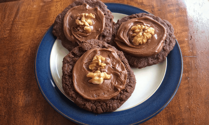 Three chocolate cookies with chocolate frosting and walnut halves on top, arranged on a blue-rimmed plate on a wooden table.
