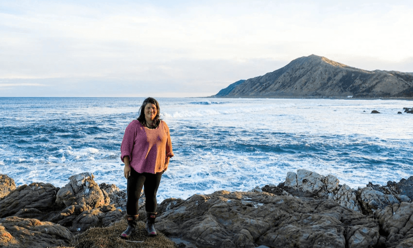 a woman in black tights and a pink long sleeve t-shirt stands on a rocky coast with the ocean in the background