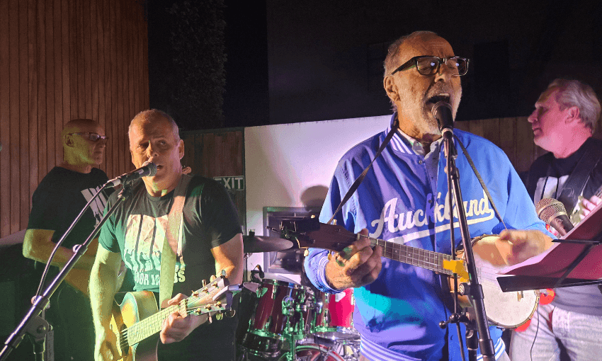 Four older men perform music on stage at night. Three play guitars while singing into microphones, and one plays drums in the background. The man in front wears glasses and a blue "Auckland" jacket.