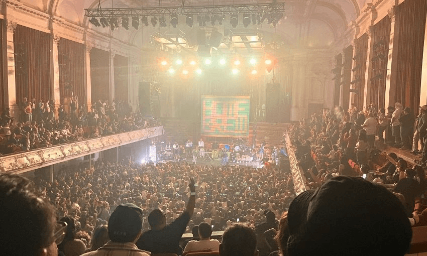 A large crowd fills a grand, ornate concert hall with balconies, watching a brightly lit stage where musicians perform. Warm lights and a colorful screen create a lively atmosphere.