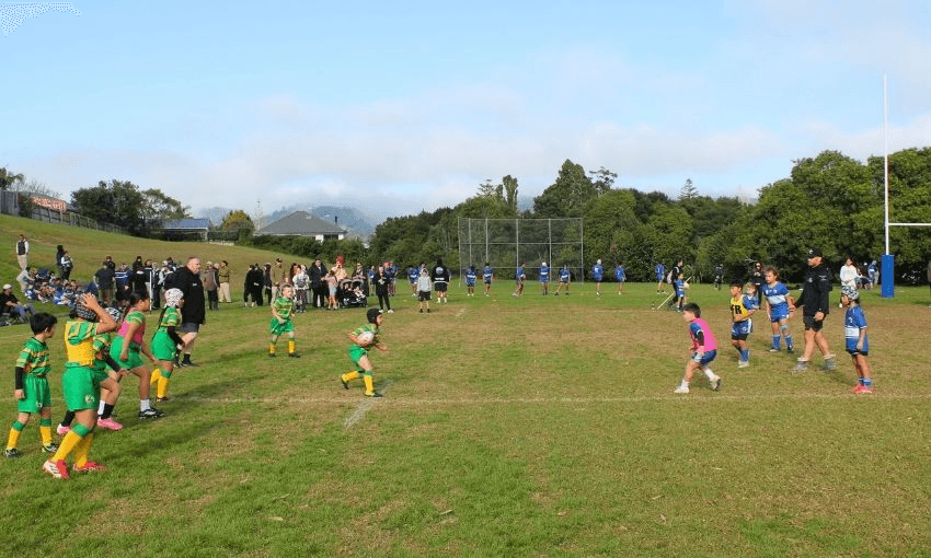 Children play a rugby league match on a grassy field, with one team in green and yellow and the other in blue and white. Spectators and coaches watch from the sidelines under a partly cloudy sky.