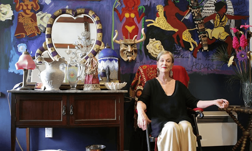 an older woman wearing a loose black top and cream skirt sits in an eclectic living room painted purple and filled with art on the walls