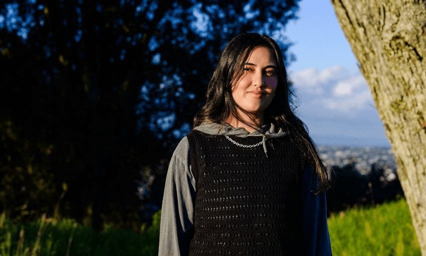 A young person with long dark hair, wearing a gray hoodie and black vest, stands outdoors near a tree, smiling, with greenery and a blurred cityscape in the background.