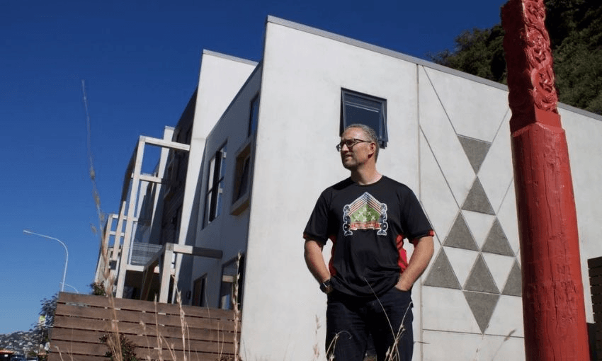 A person stands outside a modern white building with geometric designs, wearing glasses and a black T-shirt with a colorful design. A carved red post stands nearby under a clear blue sky.