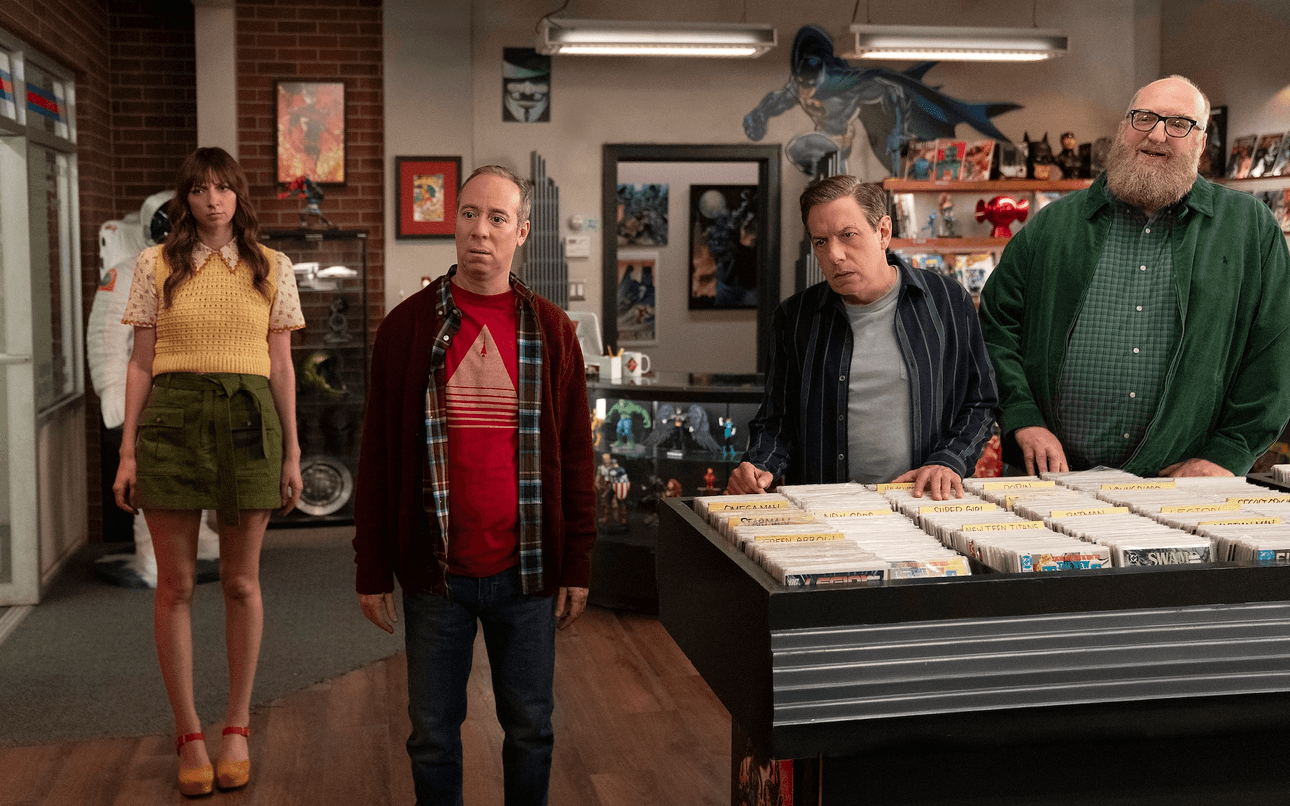 three men in plaid shirts, t-shirts and jeans stand next to a woman with long brown hair and a skirt. They are all staring intently at something off-camera and are standing in a comic book store