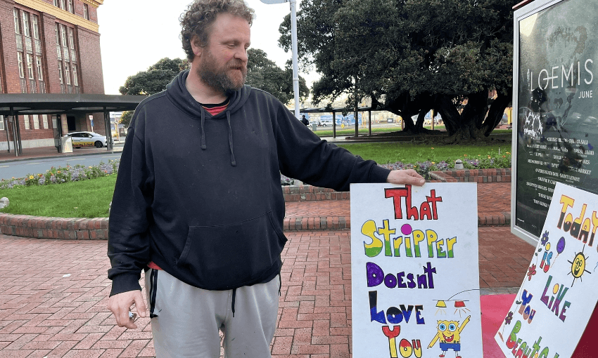 Simon standing next to a sign which reads: “that stripper doesn’t love you”.