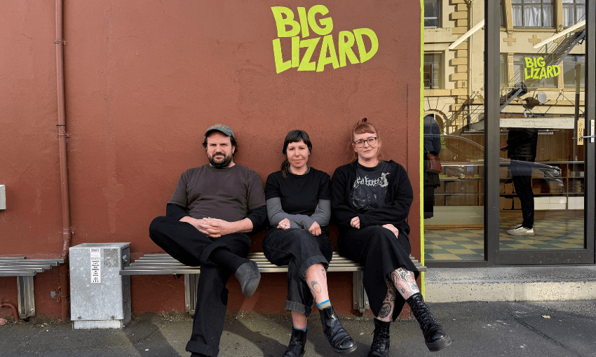 a man and two women sit on a wooden bench outside a shopfront that says "big lizard" in green writing