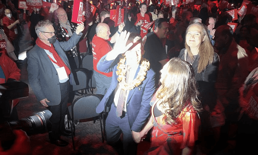 a crowded conference with people wearing red and a man in a suit and tie waving as a large camera flash goes off