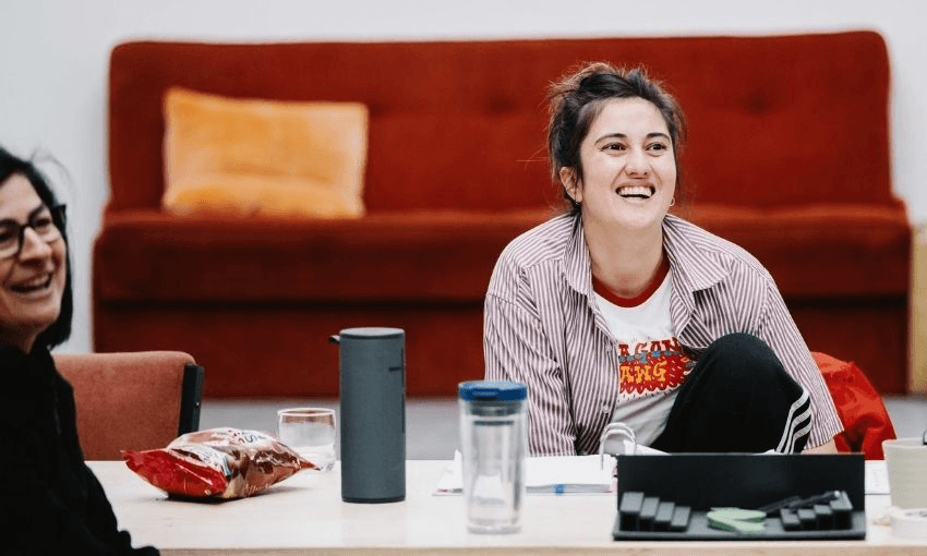 Two people sit at a table, smiling and laughing. One wears a striped shirt and sits with a knee up; snacks, drinks, and papers are on the table. A red couch and orange pillow are in the background.