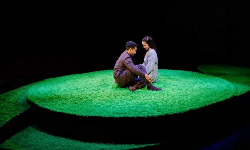 Two people sit closely together on a circular, bright green stage that resembles grass. They face each other, holding hands, and appear to be deep in conversation under dramatic lighting.
