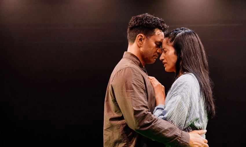A man and a woman stand close together on a dimly lit stage, gently touching foreheads and holding each other tenderly, conveying a moment of deep emotion and connection.